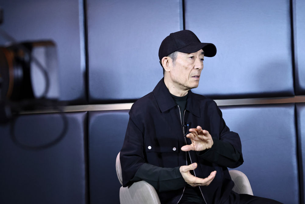 Zhang Yimou, siyah şapka ve kıyafetlerle bir röportaj sırasında koltukta oturuyor.