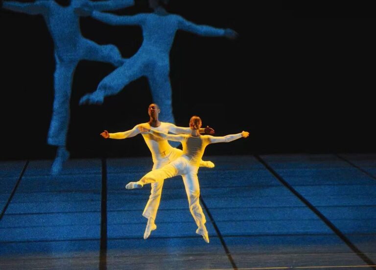 Mavi ışıklı sahnede iki dansçı, arkadaki siyah-beyaz projeksiyon önünde performans sergiliyor