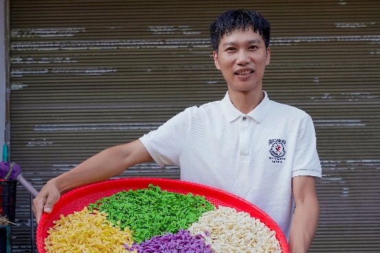 Bir adam, renkli sebzelerle süslenmiş büyük bir noodle tepsisini dükkân önünde taşıyor.